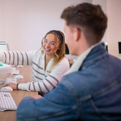 A desk with two people working on computers. One person is pointing to a computer screen while turning to face the other person.