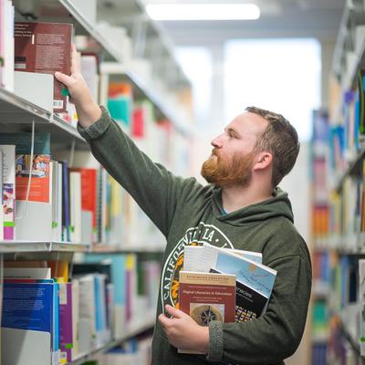 A young man with a beard reaches for a book on a library shelf, while holding several other books underneath his arm.