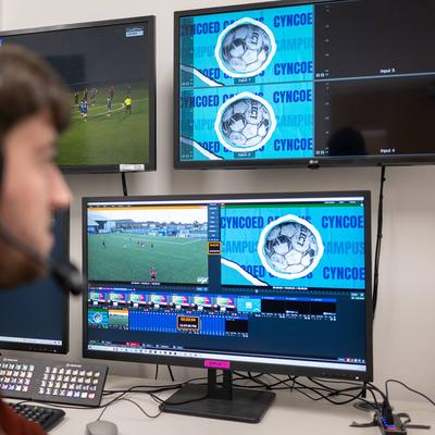 A person in headphones watches a football match on several monitors.