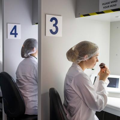 A row of people in white lab coats and hairnets sit at numbered stations. Each person is engaging in a sensory evaluation, with one person smelling or tasting from a small cake under a hooded light. The stations are numbered 3 to 8.