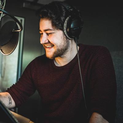 A man wearing headphones smiles while sitting in front of a microphone in a recording studio. He holds a pen and appears to be working or recording audio. The setting is dimly lit, with a lamp illuminating his workspace.