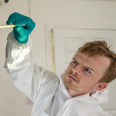 A person wearing a protective suit and teal gloves analyses fingerprints on a reflective surface, marked with green evidence tags.