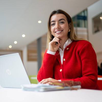 A person is smiling while sitting at a table with a laptop in front of them. They are in a bright, modern setting.