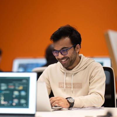 A student wearing a white hoodie and glasses is focused on his laptop while working at a desk.