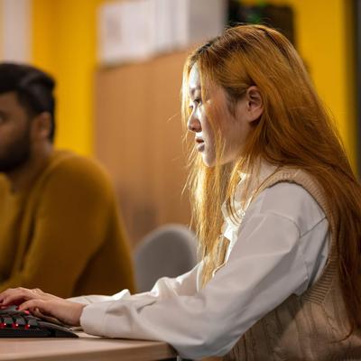A person working intently at a computer workstation. In the background another person also works at a computer.