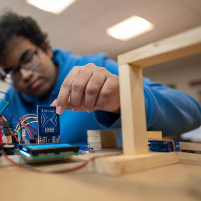 A person in a blue hoodie works with electronic components and circuit boards, placing a small blue board among connecting wires on a table. The background is blurred and the lighting is bright.