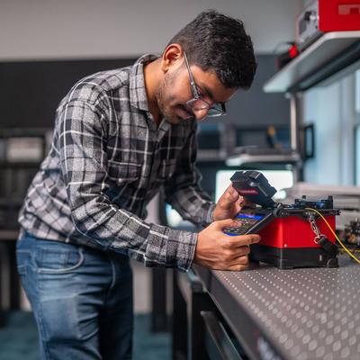 A student pressing the touchscreen of a small electronic device. In the background are other devices and computer monitors.