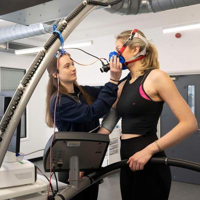 A person attaches a breathing mask to another person, who is standing on a treadmill. In front of them are computer monitors.