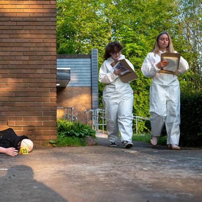 Two students wearing white overalls walk through a simulated crime scene. On the ground is a mannequin with a yellow evidence marker next to it.