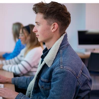 Four individuals working on desktop computers in a bright, modern workspace. The focus is on a young man in a denim jacket, working on a computer displaying a colourful SWOT analysis graphic.