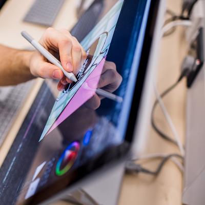 A close-up of a hand holding a stylus, drawing or writing on a digital tablet with a glowing screen. The background is blurred, creating a focused view on the tablet and the hand.