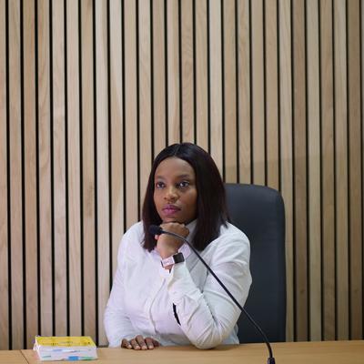 A woman seated at a wooden desk in a courtroom smiles and gestures with her hands while looking at a person sitting opposite her. The courtroom has wood panelled walls and empty leather chairs.