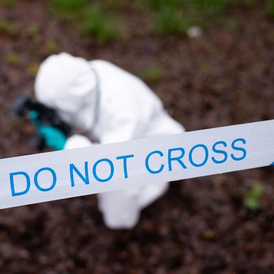 A person with a long ponytail stands outdoors behind blue and white police tape. They are wearing a dark vest with reflective strips and a badge, set against a backdrop of trees and plants.