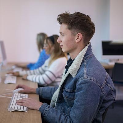A man with glasses and a mustache is smiling while using a laptop outdoors. He is wearing a green shirt and is surrounded by a blurred, sunny background of trees and greenery.