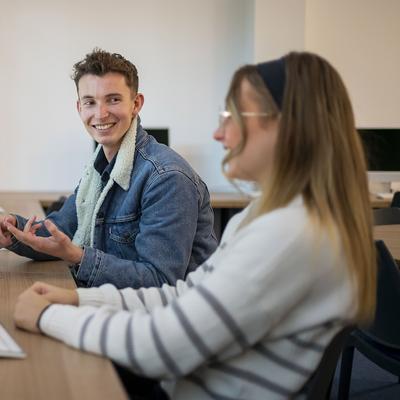 A person with long red hair is sitting at a table with a laptop and a water bottle. They are smiling and sitting in front of a large window with natural light. The background includes a yellow booth and indoor plants.