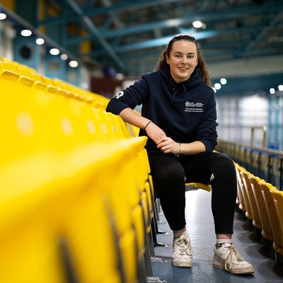 A woman with long dark hair sits on yellow bleachers in an indoor sports facility. She is wearing a black T-shirt, blue jeans, and white sneakers, and is smiling while looking to the side. The background features a running track.