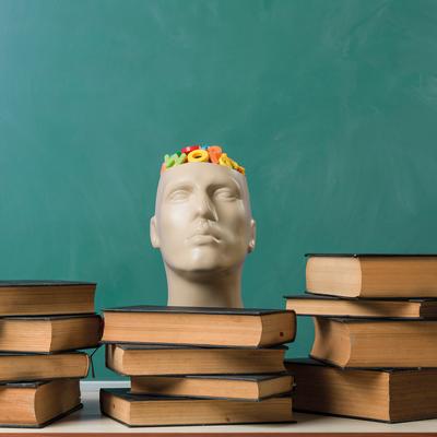 A person with curly hair is sitting at a table, smiling and writing in a notebook with a pen. They are wearing a gray sweater. The background features vertical wooden slats and a window.