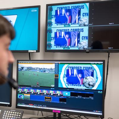 A person in headphones watches a football match on several monitors.