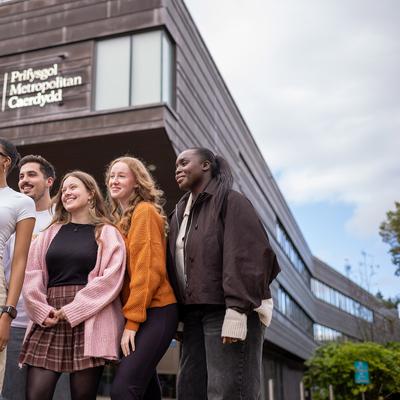 A group of four people walking and talking outdoors, holding coffee cups. Theyre dressed in casual winter clothing, with trees and modern buildings in the background under a partly cloudy sky.