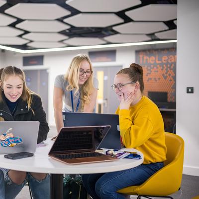 A person wearing glasses and a black t-shirt is sitting on a bright green chair, laughing. Another person with shoulder-length hair is in the foreground, slightly out of focus. The setting appears to be a casual indoor environment.