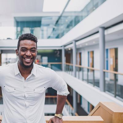 A person in a yellow jacket stands smiling in front of Cardiff Metropolitan University, with the buildings sign visible in the background against a cloudy sky.