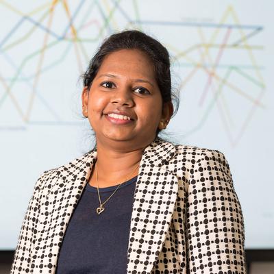 A woman with long dark hair sits on a bright green couch, smiling while using a laptop covered in various black and white stickers. She wears a black dress, and the background features a modern, minimalist office setting.