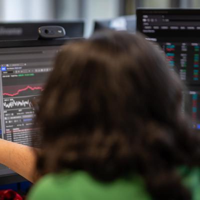A person with glasses and a mustache smiles while using a laptop outdoors. They are wearing a green jacket and sitting at a table with a mug and pens. The background is blurred, showcasing greenery and sunlight.