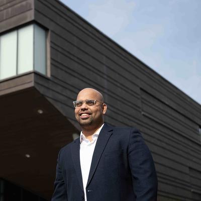 A person with glasses and a mustache, wearing a green jacket, is sitting outdoors at a table with a laptop. Theres a blue mug and a pen next to the laptop. Trees and foliage are visible in the blurred background.