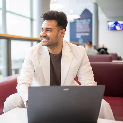 A person with long blonde hair is smiling while seated, wearing a light beige sweater. They are facing another person and have a laptop in front of them on a table. The background has a geometric pattern.