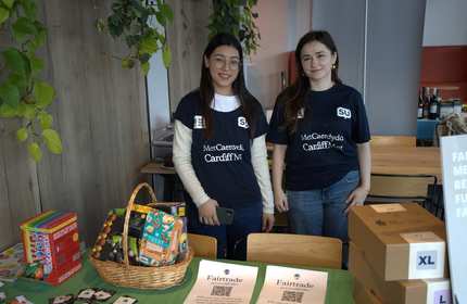 Two students stand behind a Fairtrade information stall, featuring with information sheets and Fairtrade food items
