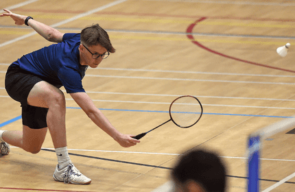 A man wearing a blue shirt and black shorts is actively playing badminton on a court.