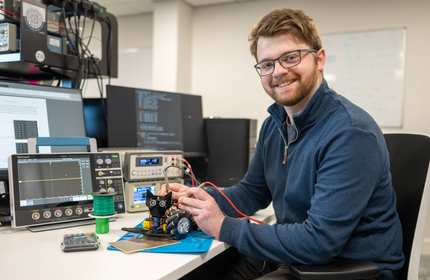 A man wearing glasses and a navy sweater is sitting at a desk with electronic equipment. Hes holding wires attached to a small device, with computer screens and measuring instruments visible around him. He is smiling at the camera.