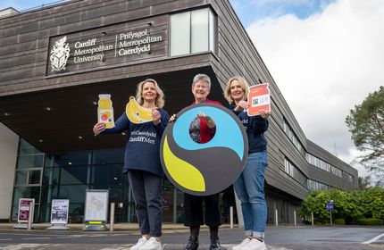 Three members of Cardiff Met staff hold Fairtrade information posters outside the Cardiff School of Management