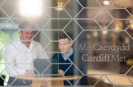Two people sit at a table working on a laptop; viewed through a glass window with Cardiff Met etched on it.