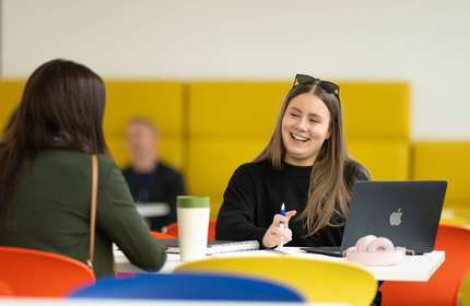 Two people sit at a table. In front of them are a laptop, notebooks and a coffee cup.