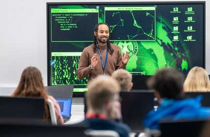A lecturer stands in front of a large screen displaying computer graphics, engaged in a presentation or discussion.