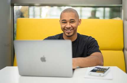 A person is smiling while sitting at a table with a laptop in front of them. They are in a bright, modern setting, with a yellow cushioned seating area behind them.