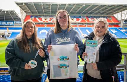 Three young women in the stands of a sports stadium hold Quick Change branded merchandise