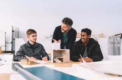 A lecturer stands between two students seated at a table with small architectural models made from card.
