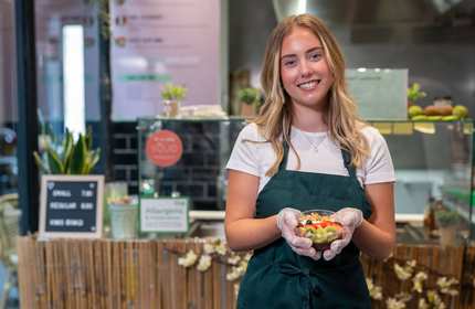 A young person wearing a green apron holds a fruit bowl while standing in front of a kitchen counter.