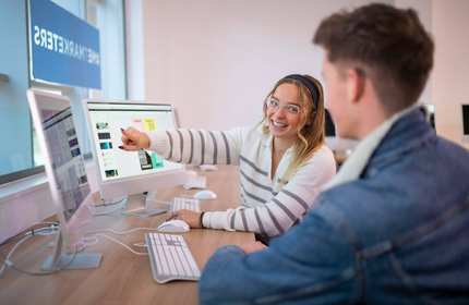 A desk with two people working on computers. One person is pointing to a computer screen while turning to face the other person.