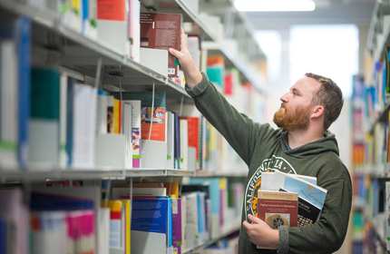 A young man with a beard reaches for a book on a library shelf, while holding several other books underneath his arm.