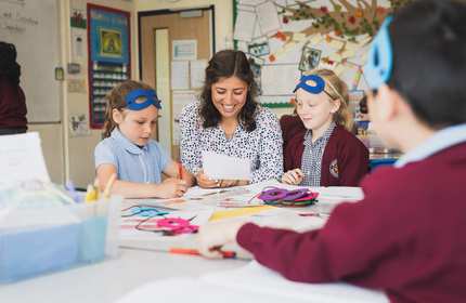 A teacher between two pupils sitting at a desk. The teacher is holding a sheet of paper.