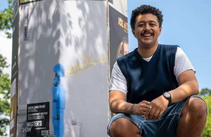 A student kneels next to a poster promoting the CSAD Master's Show.