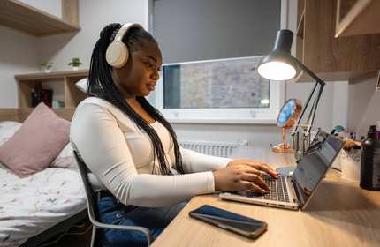 A woman wearing headphones sits at a desk, focused on her laptop while working or studying.