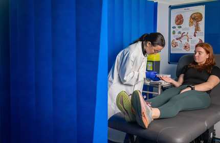 A person in a white lab coat takes a blood sample from the finger of a patient, sat on an examination bed. They are surrounded by a blue floor-to-ceiling curtain.