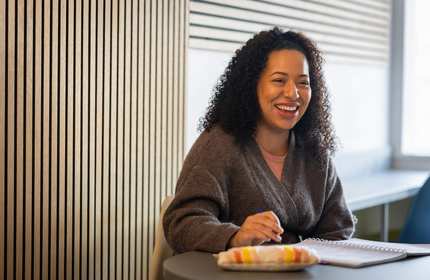 A young woman sits and smiles at a table while working with a pen and pad of paper.