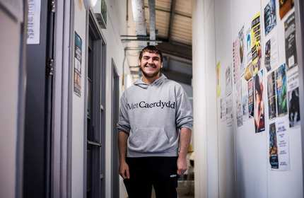 A person wearing a grey hoodie with Met Caerdydd printed on it stands in a narrow corridor.