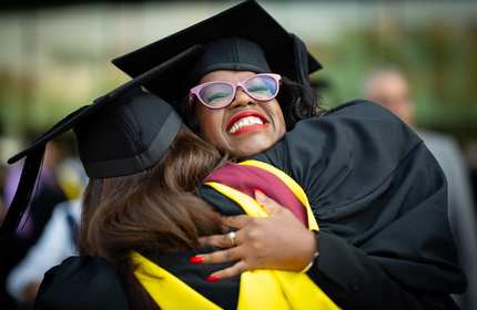 Two students in graduation caps and gowns hug each other.
