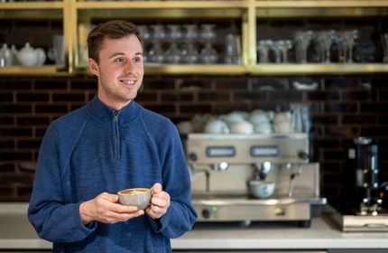 A person holding a coffee cup stands in a bar area. Behind them is a professional coffee machine.